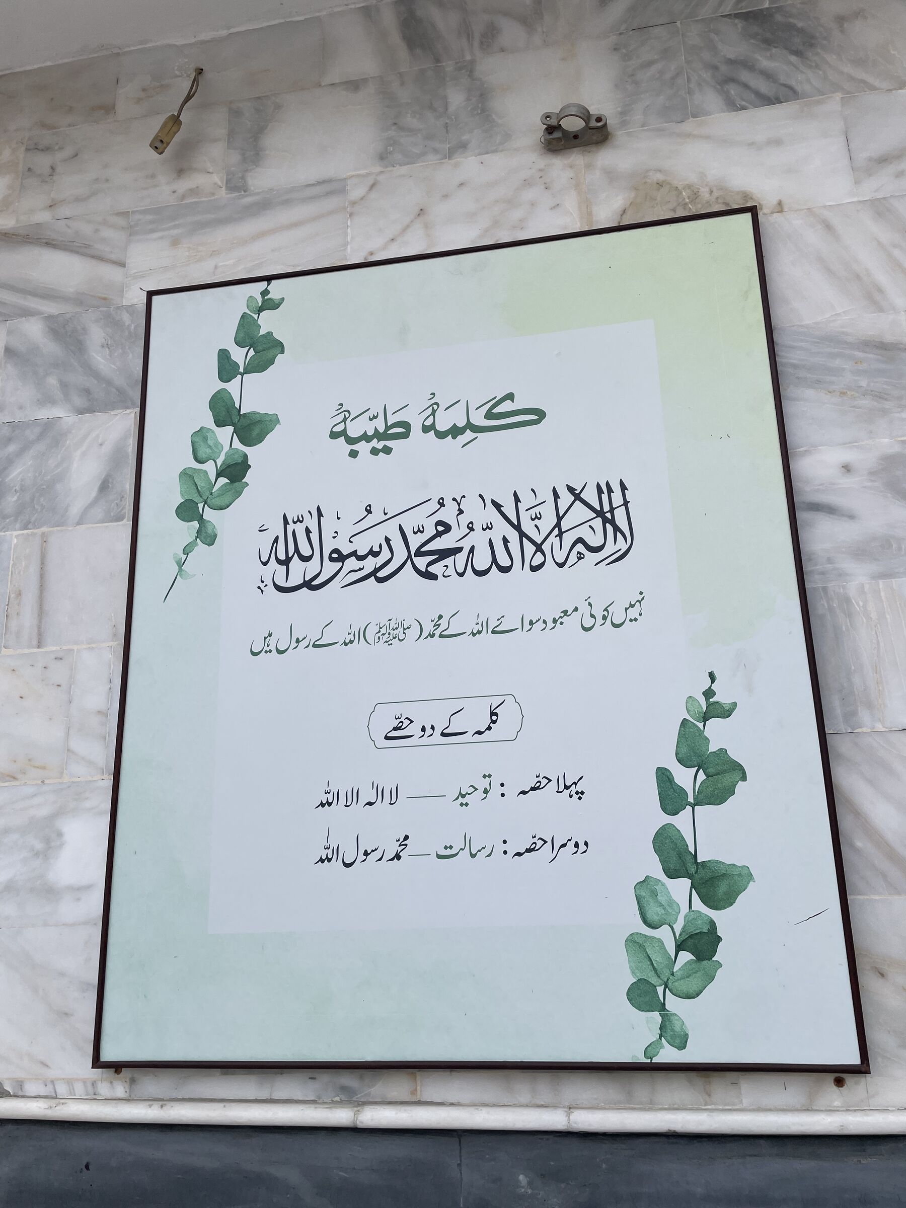 کلمہ طیبہ کا بورڈ جس کے کنارے پر سبز پتوں کی بیل بنی ہے۔