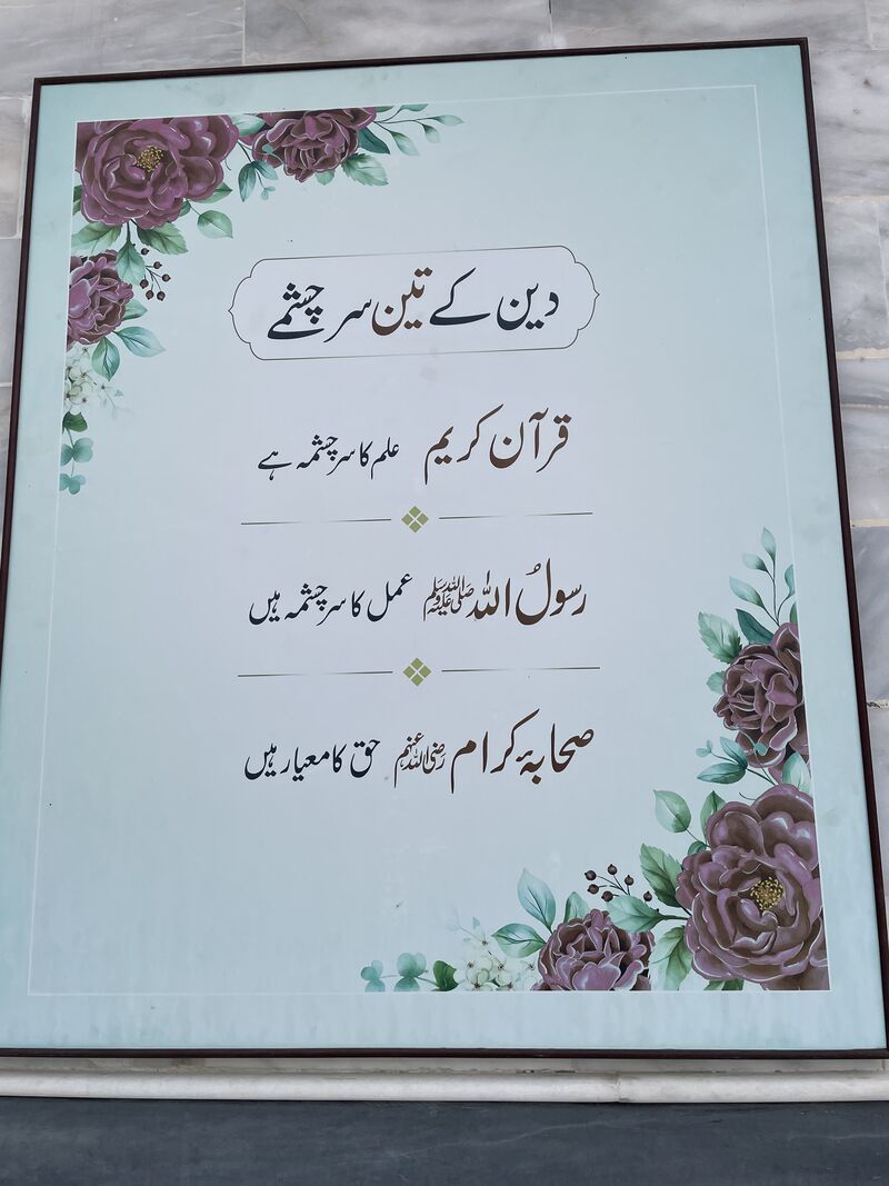 گلابی پھولوں سے مزین بورڈ جس پر دین کے تین سرچشمے درج ہیں۔